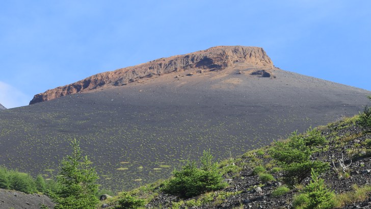 御殿場市の御殿庭から見た宝永山。赤茶色の岩の崖と黒い火山砂礫の斜面、手前に低い針葉樹