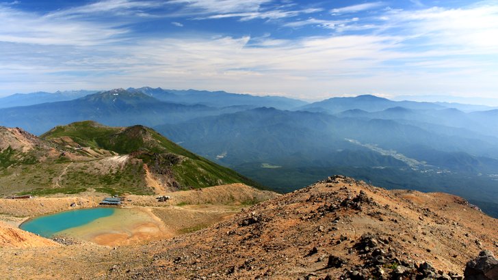 御嶽山の山頂部にある二ノ池。火山の赤茶けた岩の間に青い池があり、遠くに飛騨山脈が連なる
