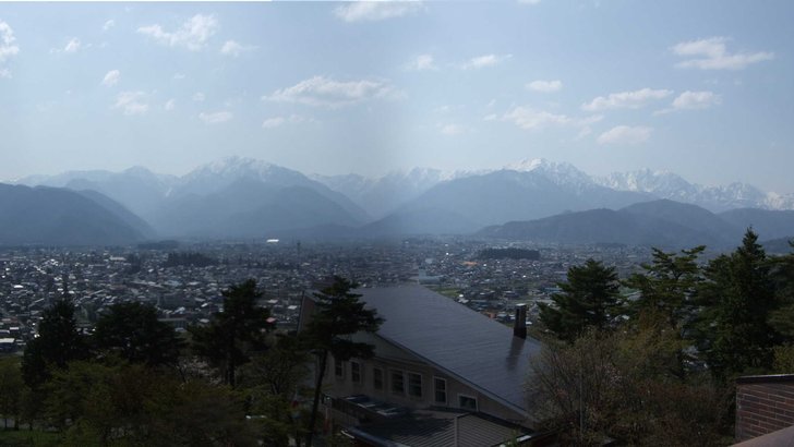 大町の市街地と北アルプスの横長パノラマ。家並みが平地いっぱいに広がり、山の斜面に雪が残る